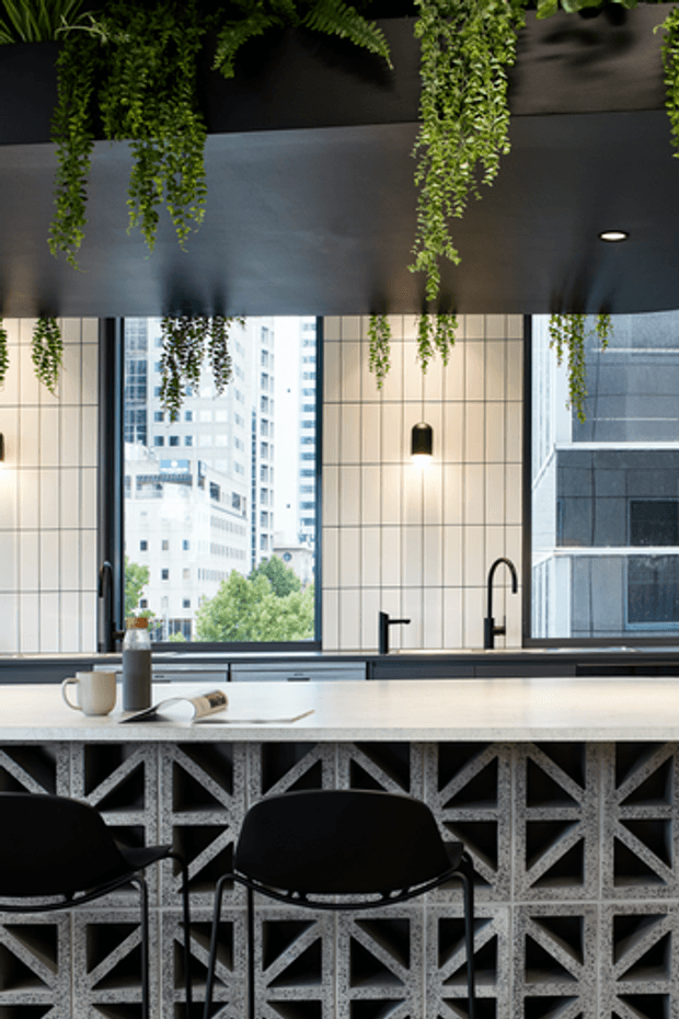 A sophisticated urban kitchen and dining counter with a pale stone benchtop, black tapware, large steel-framed windows, and a dramatic dark ceiling fitted with cascading hanging greenery. The island base features a distinctive geometric breeze-block or patterned screen, paired with black bar stools. High-rise buildings are visible through the windows, reinforcing the city setting.