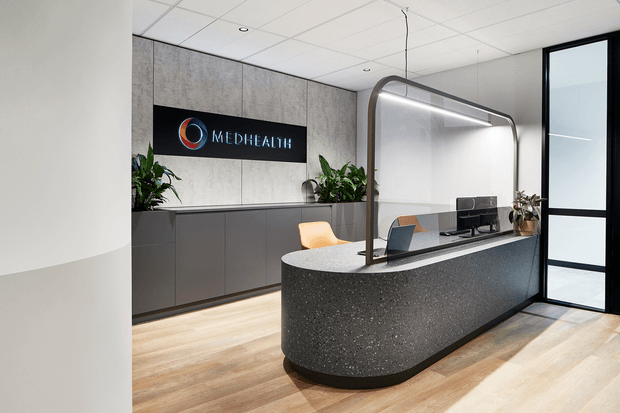 A contemporary medical reception area featuring a curved dark reception desk with an integrated protective screen, light timber-look flooring, charcoal cabinetry, a grey textured feature wall, indoor plants, and black-framed glazed doors. A MedHealth sign is mounted behind the desk.