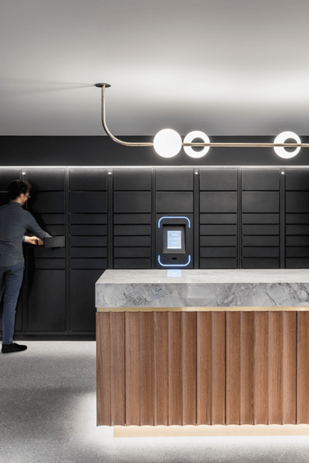 A sleek contemporary reception or service counter in a dark, highly refined interior. The foreground island combines a pale veined stone benchtop with vertically fluted mid-toned timber cladding and concealed underlighting. Behind it is a wall of matte black, paneled cabinetry or storage, with an integrated digital access or service terminal. A sculptural linear pendant with glowing circular lights hangs from the smooth ceiling, and a person stands near the rear cabinetry.