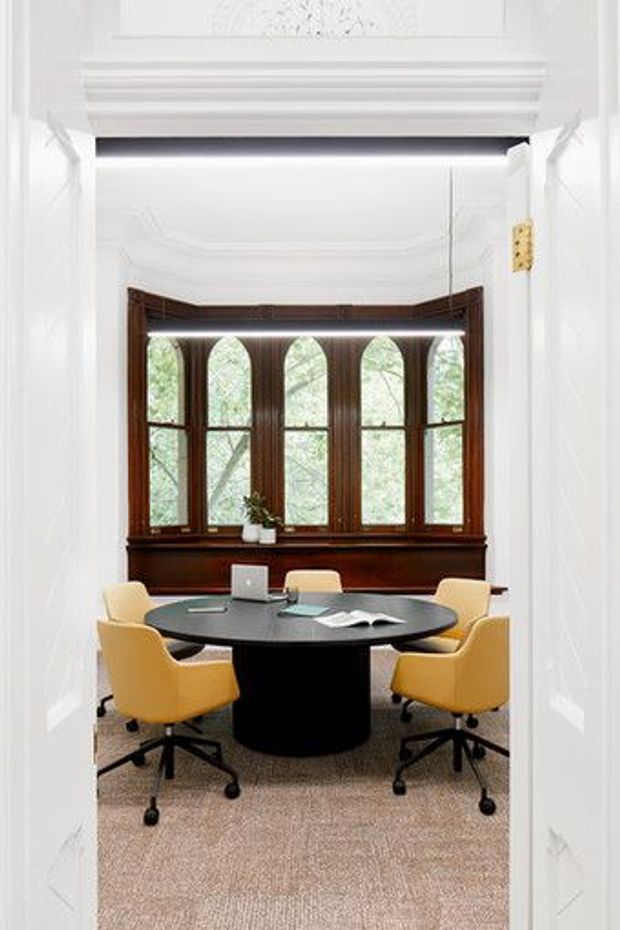 A refined meeting room viewed through open white paneled doors. The room features a dark oval conference table with a cylindrical pedestal base, four mustard-yellow upholstered swivel chairs, a laptop and papers, beige carpet, and a large dark timber bay window with pointed-arch glazing. Ornate white architectural mouldings and a decorative ceiling cornice frame the space.