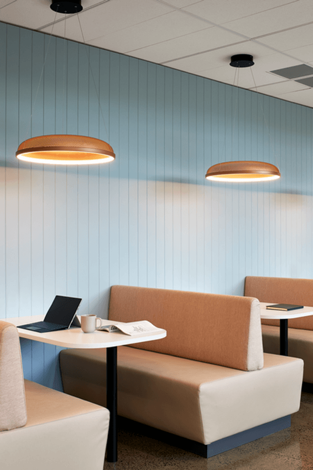 A contemporary commercial café or breakout seating area with upholstered booth seating, white tabletops, black pedestal legs, laptops and coffee cups. The space features pale blue vertical wall panelling, warm tan and cream upholstery, suspended copper-toned pendant lights, and a suspended acoustic tile ceiling.