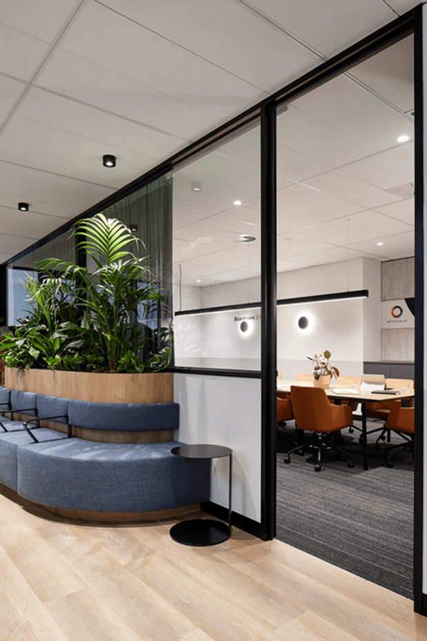 A contemporary commercial office interior with a glazed meeting room and open doorway, featuring a curved blue upholstered banquette surrounding a large built-in planter, light timber flooring, black-framed glass partitions, and a meeting table with warm orange office chairs. The space has a suspended acoustic ceiling, recessed lighting, neutral walls, and indoor greenery.