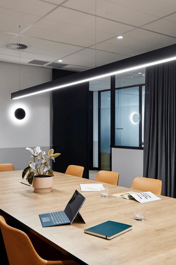 Contemporary commercial meeting room with a large light timber conference table, warm tan upholstered chairs, black-framed glazed partitions, dark curtains, and a black feature wall. A linear suspended light and circular wall light provide modern illumination, while a potted plant and office materials furnish the space.