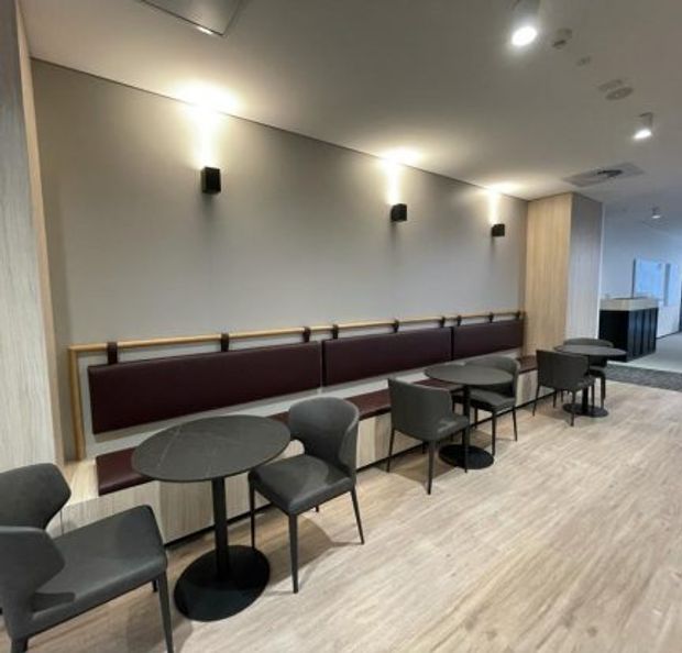A modern commercial lounge or waiting area with a long built-in banquette upholstered in deep burgundy, paired with small round black tables and dark upholstered chairs. Light timber-look wall panelling and flooring, pale walls, black wall sconces, recessed ceiling lighting, and a service counter or kitchenette area are visible in the background.