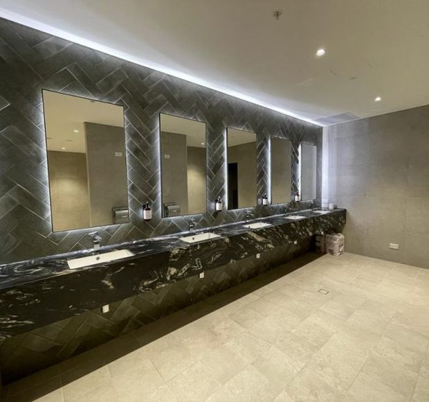 A spacious, upscale public or commercial bathroom with a long dark stone vanity and multiple integrated white basins. The feature wall is clad in dark grey geometric-patterned tiles and fitted with several tall rectangular mirrors with concealed backlighting. Pale stone-look floor and wall tiles, recessed ceiling lights, and minimalist fittings create a refined contemporary interior.