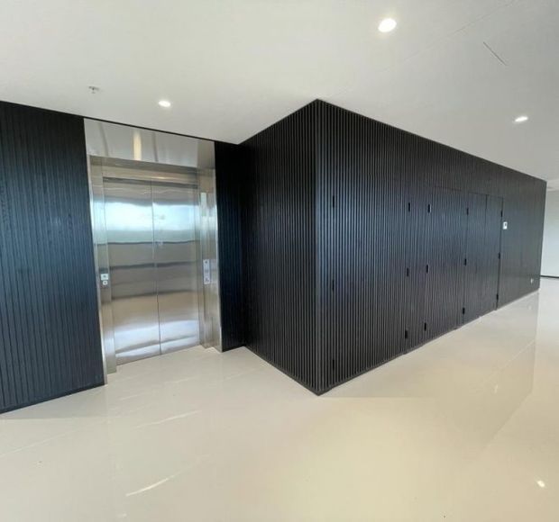 Minimalist commercial or apartment-building lift lobby with a stainless-steel elevator integrated into a dramatic black vertically fluted wall of concealed doors or cabinetry. The space has a smooth glossy pale floor, white ceiling with recessed downlights, and a clean high-contrast contemporary fit-out.