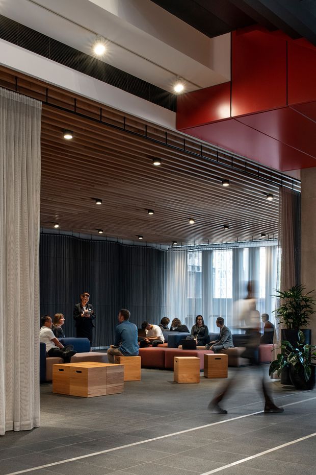 A contemporary commercial lounge or informal meeting area with multiple upholstered sofas and loose timber cube tables arranged on a large grey tiled floor. The space features a dramatic slatted timber ceiling with recessed spotlights, dark acoustic curtain walls, sheer curtains, extensive glazing, black metal detailing, and a bold red suspended ceiling or wall volume. Potted plants and blurred people add activity to the refined, hospitality-like workplace interior.