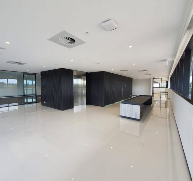 A spacious, contemporary commercial interior with a highly polished light floor, white ceiling, extensive recessed lighting, and large glazed windows. Dark vertically ribbed wall volumes and a long dark stone-topped island or reception counter create strong contrast, while glass partitions and views through the windows contribute to an open, bright workplace or showroom atmosphere.