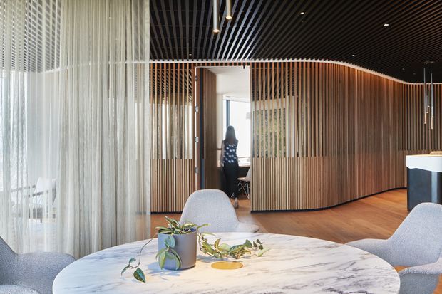 A refined contemporary interior with a curved timber slat partition and matching curved wall feature, set beneath a black linear ceiling. The foreground contains a round marble table with upholstered lounge chairs and restrained botanical styling, while sheer curtains soften the glazed edge at left. A workstation or meeting area is visible beyond the partition.