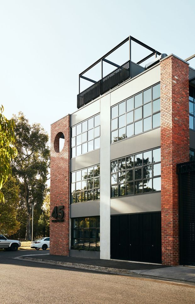 Street-facing multi-storey contemporary building with a strong red-brick facade, white rendered vertical sections, large black-framed industrial-style grid windows, and a black garage or entry door. The upper level has a rooftop terrace enclosed by a black metal balustrade and open geometric frame. An arched opening in the brickwork and bold address numerals provide distinctive facade details. Mature trees and parked vehicles appear in the surrounding street context.