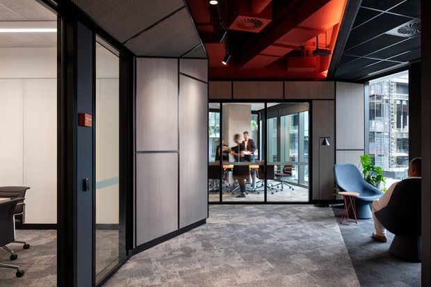 Contemporary commercial office interior with glazed meeting rooms, black-framed partitions, acoustic ceiling panels, carpeted flooring, and a dramatic exposed red services ceiling. The central view looks toward a furnished meeting room, with lounge seating and a plant in the foreground.