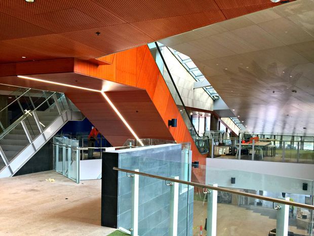 Interior view of a large contemporary commercial atrium with intersecting staircases, glass balustrades, polished stone surfaces, exposed geometric circulation elements, and dramatic orange timber-lined ceiling and wall planes. A long linear skylight brings daylight into the expansive multi-level space.