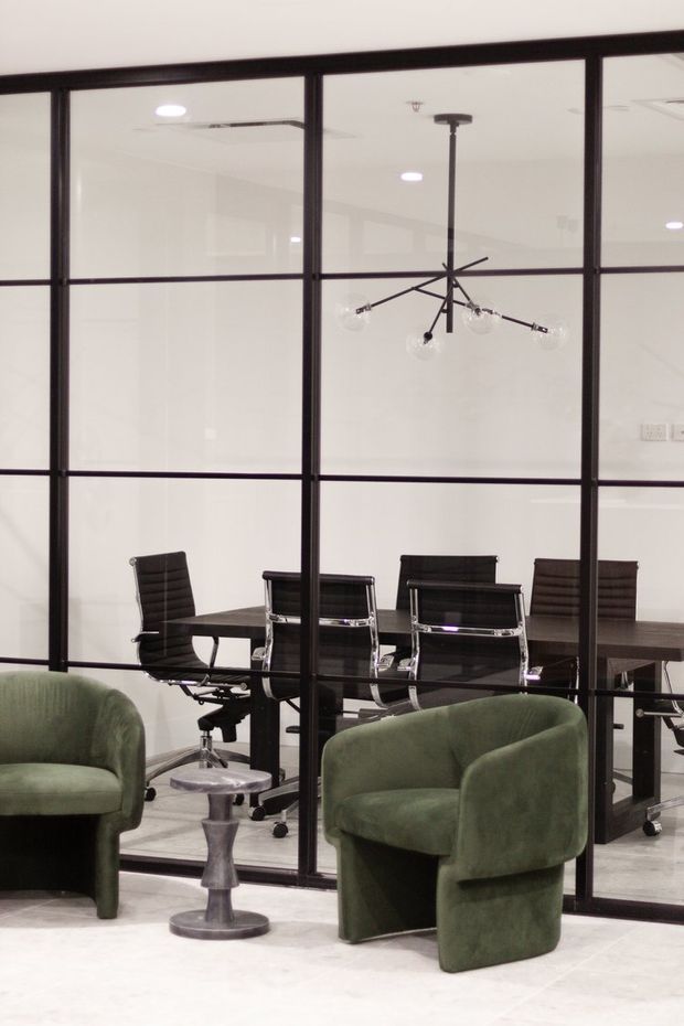 A contemporary office or meeting area viewed through a black-framed glass partition. A dark rectangular meeting table with black office chairs occupies the enclosed room, while two deep green upholstered lounge chairs and a small metallic side table sit in the foreground. A sculptural black multi-arm pendant light hangs above the table. The space has white walls and ceiling, pale flooring, and strong black framing details.