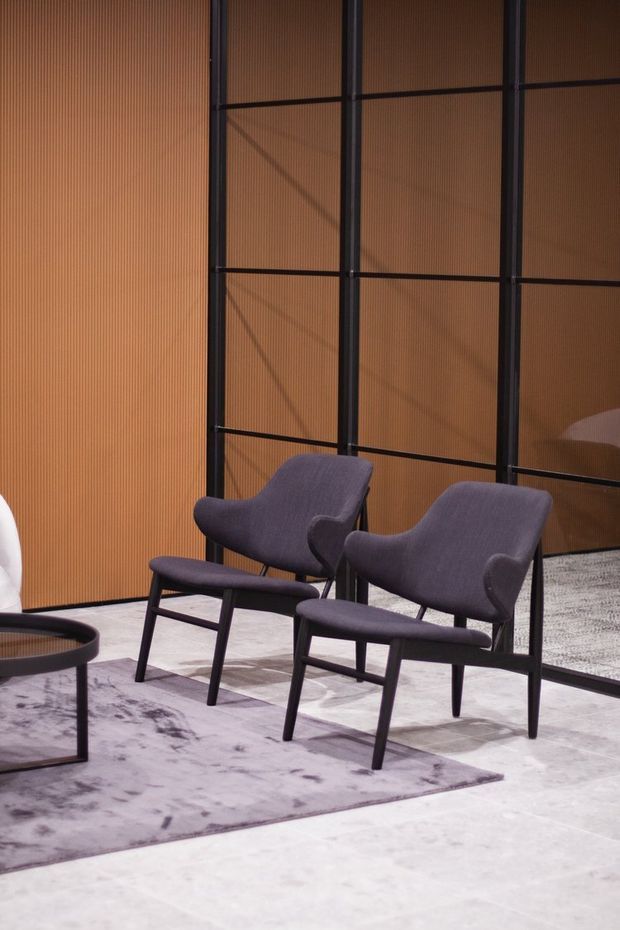 A refined seating vignette featuring two sculptural upholstered lounge chairs in dark charcoal fabric with black timber or metal frames, arranged on a muted patterned rug. A low round coffee table is partially visible at left. The setting has warm vertical timber slat wall panelling and a black-framed glazed or screened partition, creating a sophisticated contemporary interior.