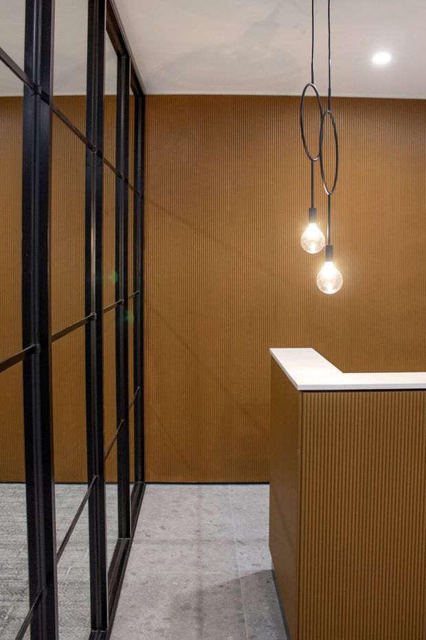 A refined contemporary interior featuring a reception or service counter with a white top and vertically ribbed warm timber-toned wall and counter fronts. Black metal-framed glazed partitions line the left side, while two suspended loop pendant lights provide a sculptural focal point. The floor is finished in pale grey stone or concrete-look material, creating a warm, modern commercial atmosphere.