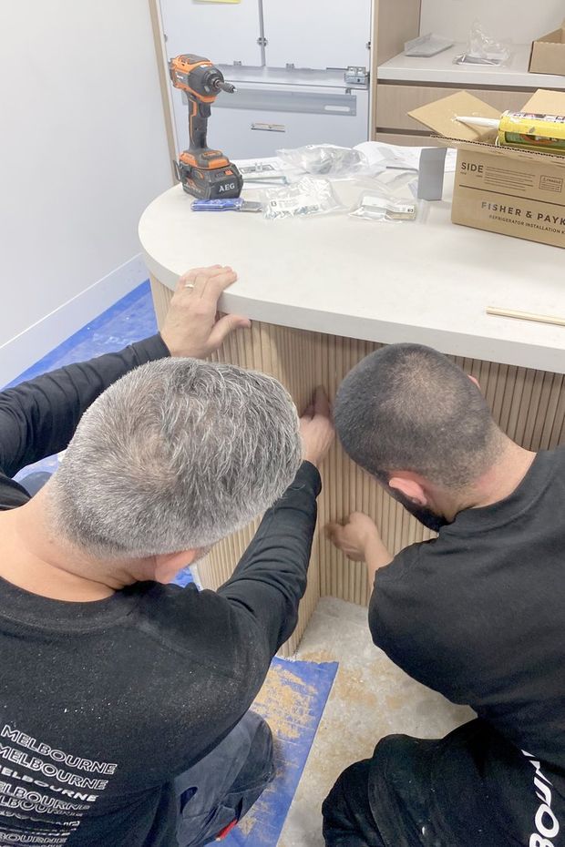 A construction or installation scene inside a workshop or kitchen-like fit-out area. Two tradespeople are working on a curved white benchtop and vertically fluted timber-look cabinetry. Tools, packaging, a drill, and unfinished flooring protection are visible around the work area.