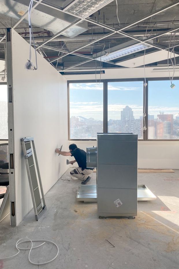 An unfinished high-rise commercial interior under construction, with exposed concrete ceiling, suspended ceiling grid, visible wiring and ductwork, partially installed white partition walls, large dark-framed windows, and construction equipment. A worker is installing or finishing a wall, and a bright city skyline is visible through the glazing.