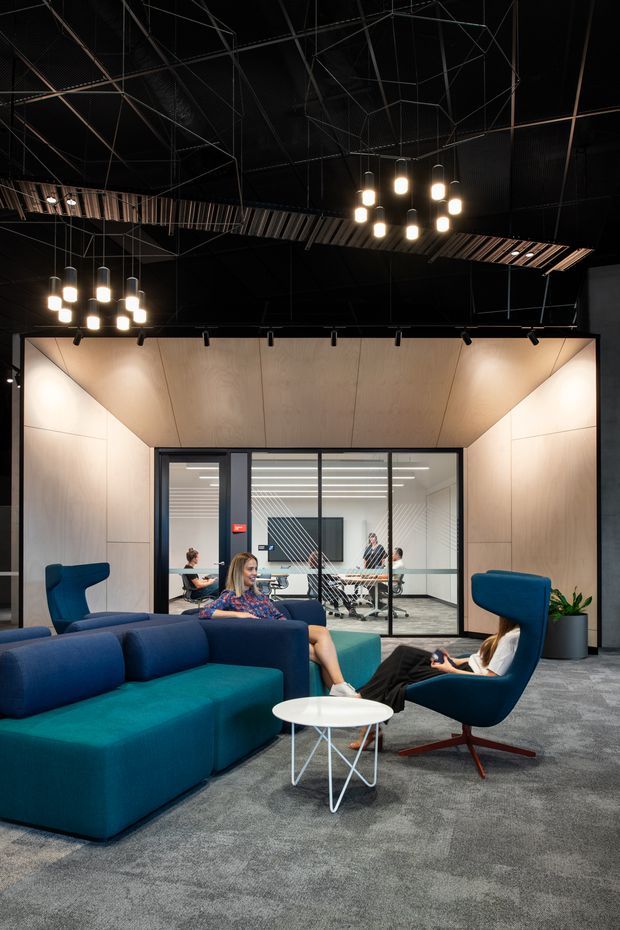 A dark, dramatic contemporary commercial interior showing a relaxed office lounge and breakout area. Deep teal and navy upholstered sofas and armchairs surround small white round tables on grey carpet. Behind the lounge is a glazed meeting room with desks and office chairs. The space features warm light timber wall and ceiling panels, black exposed ceiling structure, track lighting, and clustered pendant lights.