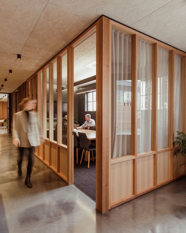 A warm contemporary commercial interior showing a timber-framed glazed meeting room opening onto a polished concrete corridor. The enclosure features vertical timber mullions, clear glazing, sheer curtains, and lower timber panels. Inside, a meeting table and chairs are visible, while a blurred person walks through the corridor. The space has a timber-lined ceiling, dark carpet in the meeting room, and soft natural light.