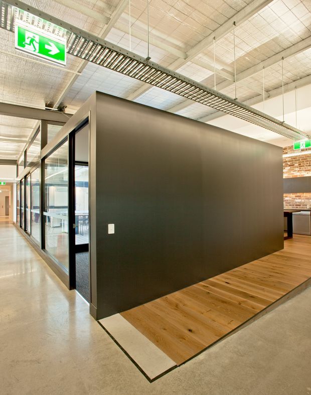 Modern commercial office interior with a large dark partition wall, glazed meeting-room frontage, polished concrete flooring, and a contrasting timber floor zone. The exposed industrial-style ceiling has suspended linear lighting and visible services, while brickwork and workspaces are visible in the background. A green emergency exit sign is suspended overhead.