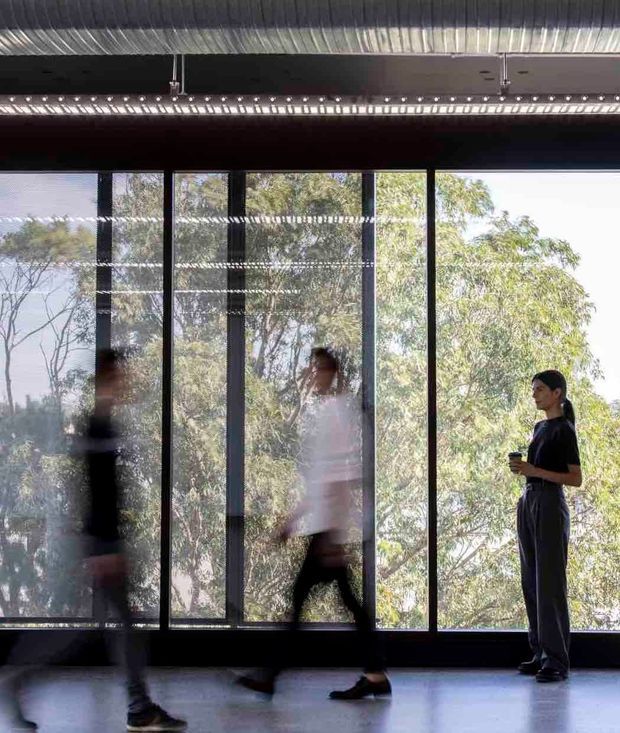 A contemporary interior corridor or open workspace with full-height black-framed glazing overlooking trees. Several people are walking through the space, while one person stands holding a cup. The room has a dark exposed ceiling with large cylindrical metal ductwork, linear suspended lighting, and a smooth polished grey floor, creating an industrial architectural atmosphere.