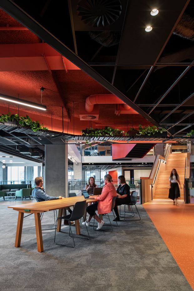 A contemporary open-plan commercial interior, likely an office or collaborative workplace. The space features exposed black ceiling services and structure, dramatic red-orange textured ceiling and wall surfaces, linear pendant lighting, indoor planting integrated along a mezzanine edge, glass meeting rooms, concrete columns, and a prominent timber staircase with orange flooring. Several people are seated at a communal timber worktable, with lounge seating visible in the background.