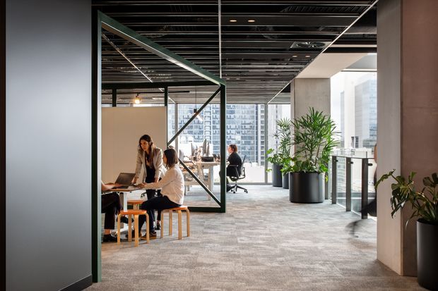 Contemporary open-plan commercial office interior with carpeted flooring, exposed dark ceiling services, extensive floor-to-ceiling glazing, indoor plants, and collaborative work areas. Employees are seated at desks and stools, with city high-rise buildings visible through the windows. A dark metal-framed glazed partition creates a strong geometric feature in the foreground.