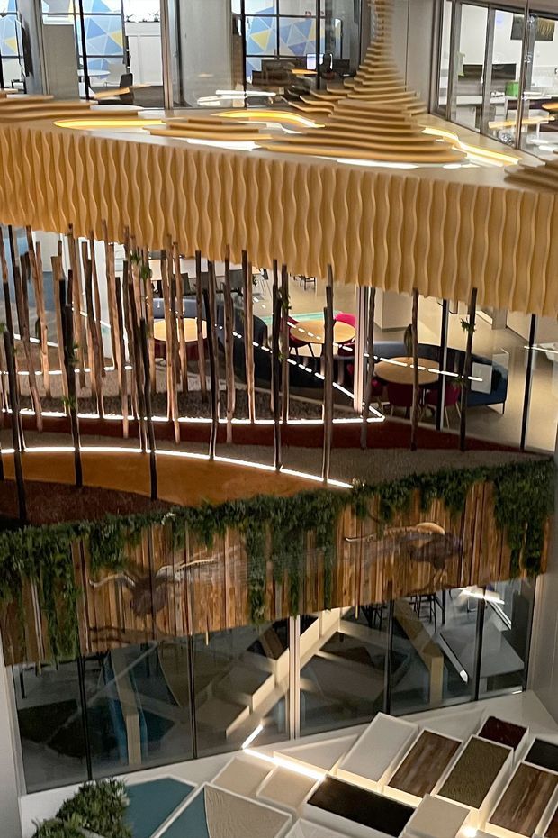 A vertically oriented view into a multi-level commercial interior atrium, featuring extensive glass balustrades and partitions, sculptural suspended timber-toned wave panels, layered illuminated ceiling forms, a planted feature wall, and seating areas visible below. The space has a contemporary, highly designed hospitality or workplace environment with strong geometric lines and decorative architectural detailing.