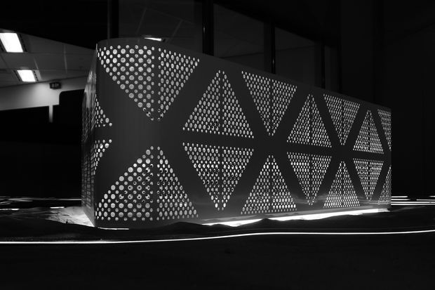A dramatic black-and-white architectural interior featuring a large curved perforated metal screen or partition. The screen is decorated with repeated triangular geometric patterns made from circular cut-outs and is illuminated from behind, creating a strong graphic glow. The surrounding space is dark and appears commercial or exhibition-like, with linear lighting and reflective dark surfaces.