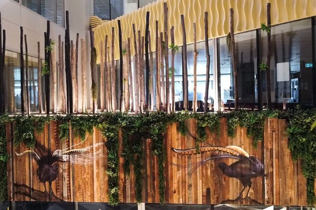 A contemporary commercial interior feature installation viewed across a decorative timber-clad partition. Tall natural branches form a vertical screen in front of glazing, with lush planted greenery along the top and a sculptural yellow undulating ceiling feature above. The timber partition includes large bird-like decorative artwork, while a reception or dining area is visible behind the screen.