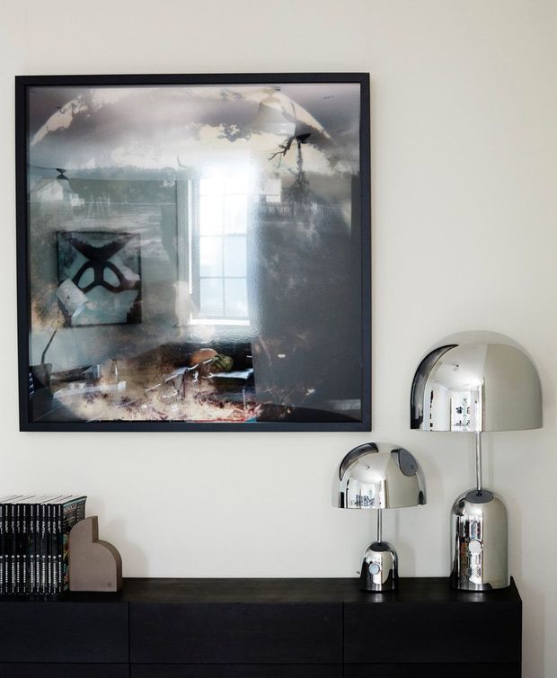 A refined interior vignette featuring a dark built-in sideboard against an off-white wall, topped with reflective chrome dome table lamps and a small stack of books. A large dark-framed abstract artwork hangs above, with its reflective surface showing parts of the room. The composition has a clean, curated contemporary feel with strong black, white, and metallic contrasts.
