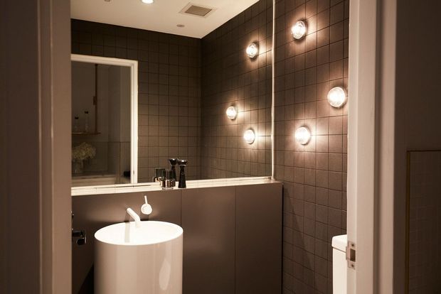 A compact contemporary bathroom viewed through an open doorway, featuring dark square wall tiles, a white pedestal basin with a wall-mounted tap, a large mirror, and multiple round wall sconces. A toilet is partially visible at the right, with warm, moody lighting and dark neutral finishes.