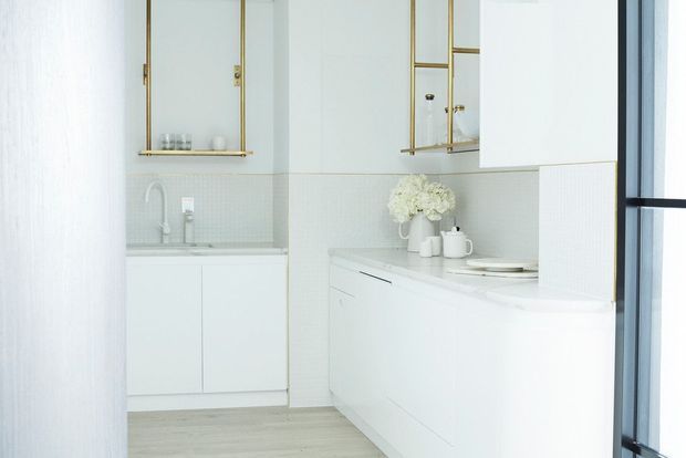 Bright, compact contemporary kitchen with white flat-panel cabinetry, a pale tiled splashback, light timber flooring, and a curved white peninsula or island. Brass-framed glass-front wall cabinets add a refined accent, while large black-framed glazing is visible at the right. The space has a clean, airy, minimalist appearance.
