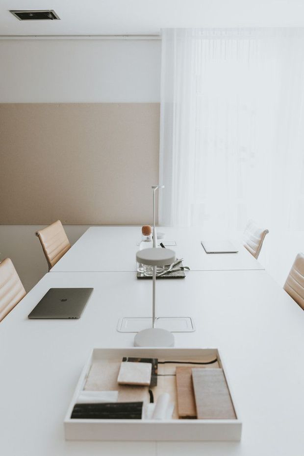 A bright, minimalist meeting or dining room with a long white rectangular table, integrated central power modules, modern desk lamps, tan upholstered chairs, a laptop, and neatly arranged design samples. Sheer white curtains cover large glazing, while a beige acoustic or textured wall panel provides contrast against the white ceiling and furniture.