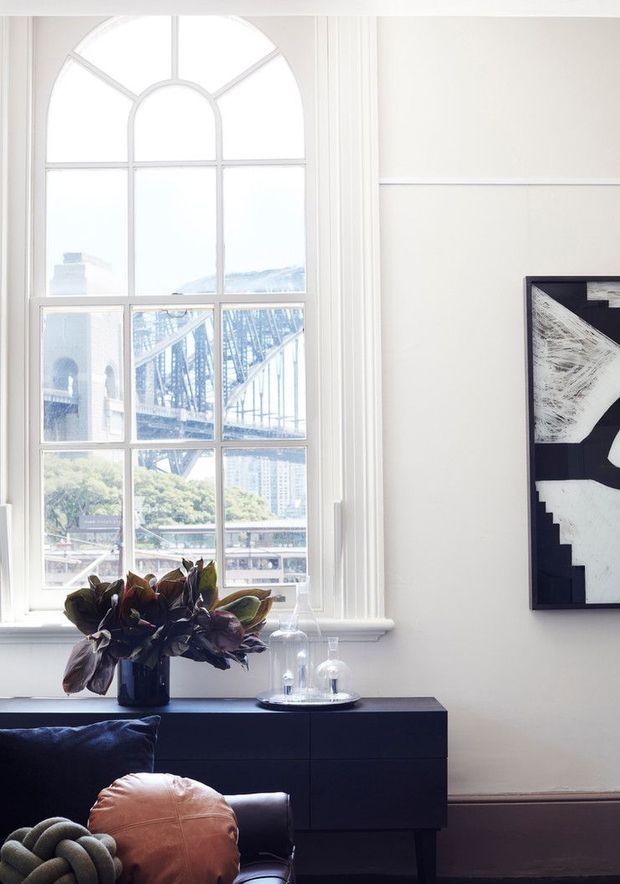 Bright, refined living room interior featuring a tall arched multi-pane window with decorative white trim and views of an urban bridge and buildings beyond. A dark low-profile sideboard sits beneath the window, styled with leafy foliage, glass cloches and decorative objects. A black leather sofa with a tan cushion occupies the foreground, while a large monochrome abstract artwork hangs on the pale wall.