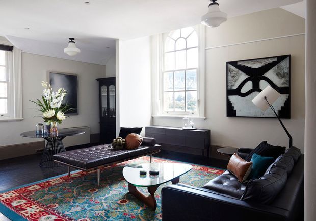 Bright, refined living room with white walls, tall arched windows, dark leather seating, a tufted chaise, black cabinetry, and a colourful patterned area rug. Abstract monochrome artwork, sculptural lighting, and a glass-topped coffee table create a sophisticated eclectic interior.