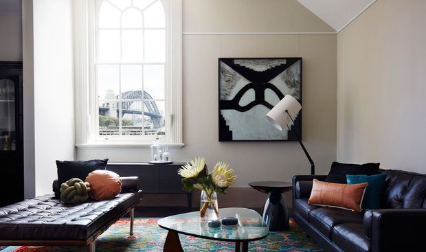 Refined living room with a high pitched ceiling, large arched-top window, dark built-in cabinetry, black leather seating, a tufted chaise, patterned area rug, glass coffee table, floor lamp, and abstract wall art. The room combines neutral architectural finishes with rich dark furniture and colourful accents.
