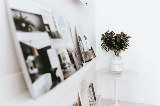 Bright minimalist interior vignette featuring a white picture ledge displaying monochrome and neutral-toned photographs, alongside a small round white pedestal table with a glass vase of leafy greenery. The space has white walls, pale flooring, and a clean, airy aesthetic.