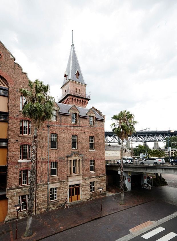 Exterior view of a historic red-brick building with a prominent pointed bell tower, arched and mullioned windows, stone detailing, and a steep slate-clad spire. Palm trees, elevated roadway structures, and an urban streetscape surround the building.