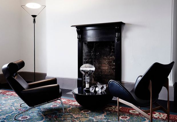A refined living room with a black-painted traditional fireplace surround and exposed brick firebox, white walls, dark trim, sculptural black leather lounge chairs, a round black coffee table, a tall floor lamp, and a richly patterned area rug. The room combines classic architectural detailing with mid-century-inspired furniture and dramatic dark accents.