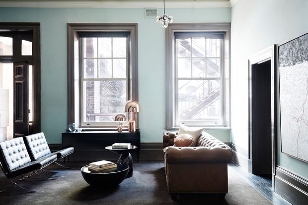 A refined living room with pale blue-green walls, dark timber window and door trim, tall sash windows, dark flooring, a tufted black leather lounge chair, brown leather sofa, black coffee table, and a slim black console with copper-toned fixtures. A large framed artwork and sculptural pendant add polished decorative accents. The room has generous ceiling height and abundant natural light.