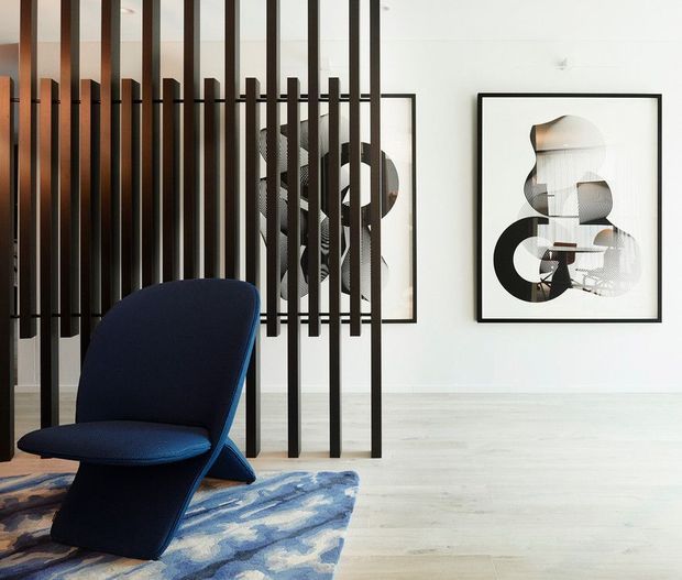 Contemporary interior featuring a sculptural navy lounge chair on a patterned rug, pale timber flooring, white walls, black-framed abstract artwork, and a striking black vertical timber-slat room divider with additional artwork visible behind it.
