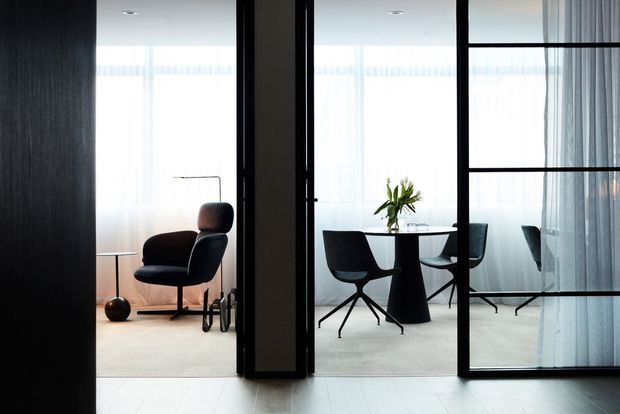 Interior view through black-framed glazed partitions into a refined open-plan lounge and dining area. A dark upholstered lounge chair and slim floor lamp sit in the sitting zone, while a round dining table with sculptural pedestal base and contemporary chairs occupies the brighter room beyond. Sheer curtains, pale flooring, dark timber or charcoal wall surfaces, and strong daylight create a restrained, elegant atmosphere.