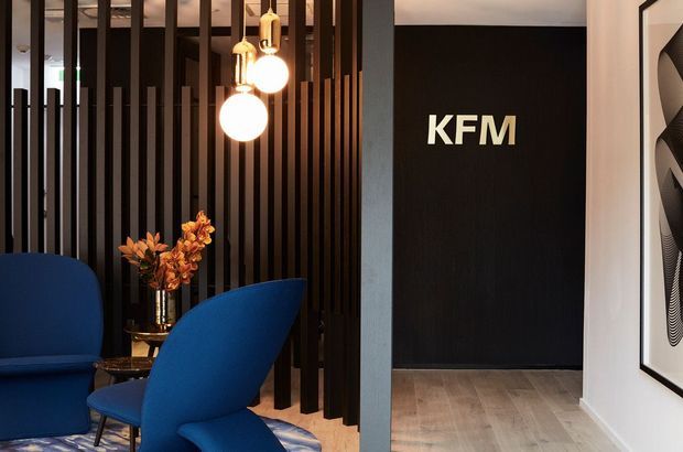 A refined commercial reception or waiting area featuring sculptural deep-blue lounge chairs, a dark vertical timber-slat partition, warm globe pendant lighting, and a black feature wall marked “KFM.” Light timber flooring, a floral arrangement, and framed artwork add warmth and visual interest.