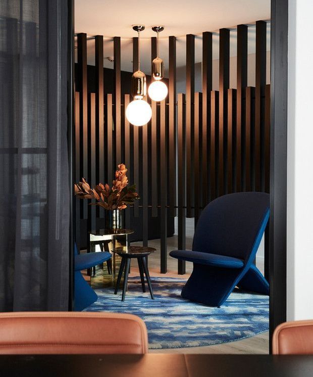 A dramatic, contemporary lounge or reception area viewed through a doorway, featuring dark vertical timber or metal screening, sculptural navy lounge chairs, a blue patterned rug, warm pendant globe lights, and a small round side table with dried botanical decor. Partial tan upholstered seating and a dark table appear in the foreground.