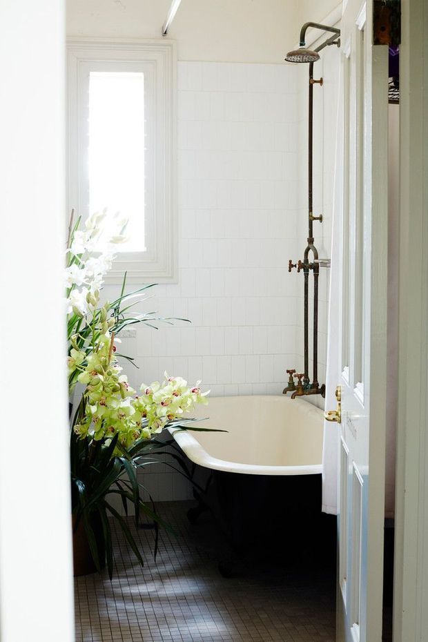 A narrow, elegant bathroom viewed through an open white paneled door. The room features a freestanding black-and-white clawfoot-style bathtub, a vintage exposed bronze shower fixture, white rectangular wall tiles, small dark floor tiles, a tall window, and a large floral arrangement.