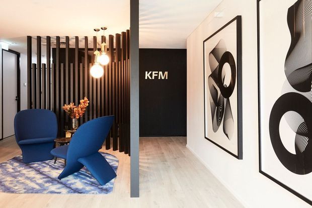 A stylish contemporary interior featuring a compact lounge or reception seating area separated from a hallway by a vertical dark timber slat screen. Two sculptural blue lounge chairs sit on a patterned rug beside a small round table, with warm pendant lighting overhead. The adjacent white hallway has pale timber flooring, bold black-and-white framed abstract artwork, and a dark feature wall marked “KFM.”