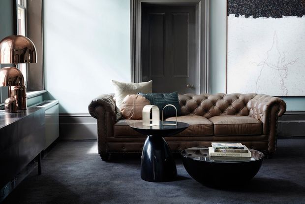 A refined living room with a deep brown tufted Chesterfield-style sofa, dark carpet, black sideboard, sculptural black coffee tables, and polished copper dome lamps. Pale blue-grey walls, dark timber trim, a black paneled door, and a large abstract artwork create a sophisticated, moody interior.