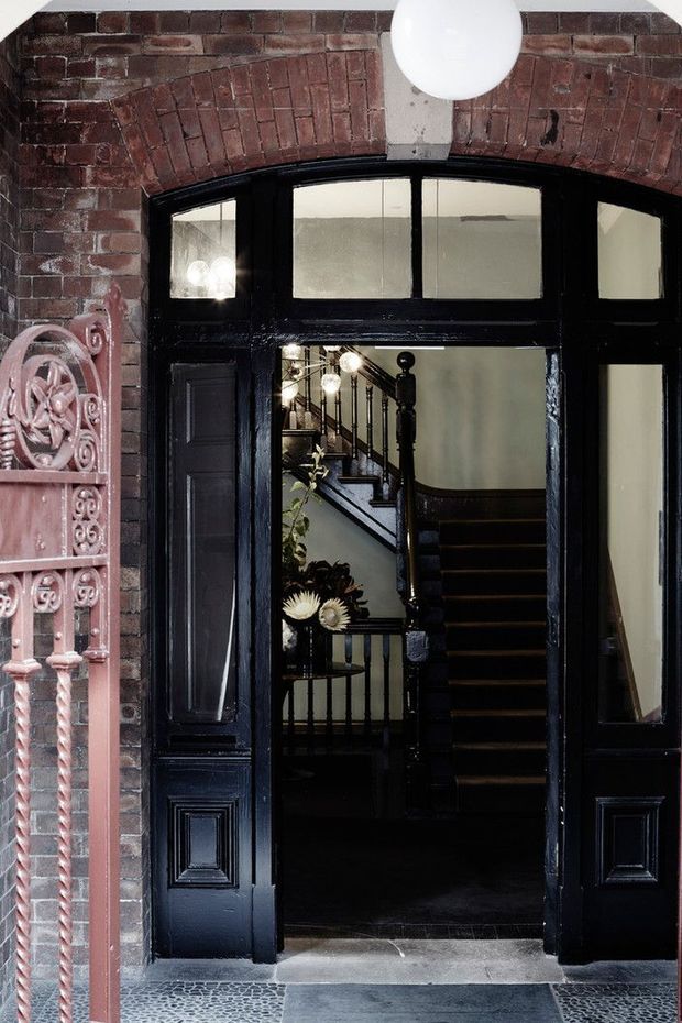 A dramatic heritage-style entryway viewed from outside, featuring a black-painted timber entrance with sidelights and fanlight glazing, set within an ornate red brick arched facade. A dark stairway and hallway are visible beyond the doorway, with a white globe pendant light overhead and decorative metalwork at the left edge.