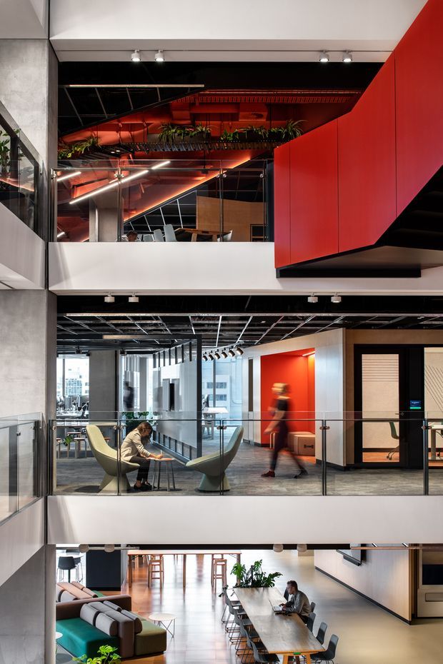 A multi-level contemporary commercial interior, likely an office atrium, viewed across open floor plates and glass balustrades. The space features exposed black ceilings and services, dramatic red architectural wall and ceiling elements, white structural bands, concrete-toned columns, timber joinery, and extensive glazing. Workspaces, lounge seating, communal tables, and people are visible across the levels, with strong vertical layering and an open void connecting the floors.